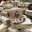 A trucker-style cap with a tan crown and camouflage-patterned brim sits on stacked bricks. The front panel features a shield-shaped embroidered logo reading 'Los Surf Shop' with a sunset and ocean design.