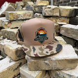 A trucker-style cap with a tan crown and camouflage-patterned brim sits on stacked bricks. The front panel features a shield-shaped embroidered logo reading 'Los Surf Shop' with a sunset and ocean design.