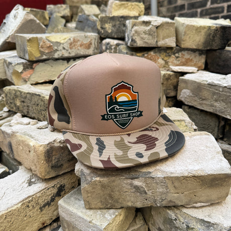 A trucker-style cap with a tan crown and camouflage-patterned brim sits on stacked bricks. The front panel features a shield-shaped embroidered logo reading 'Los Surf Shop' with a sunset and ocean design.