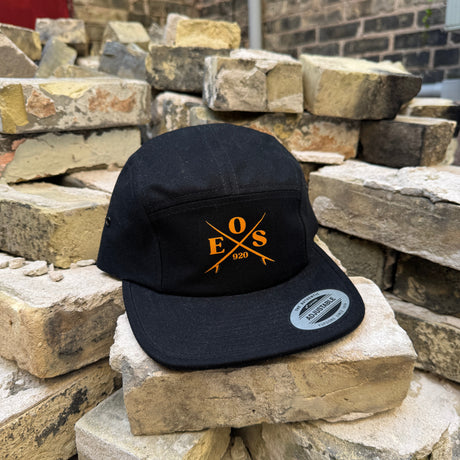 A black 5-panel cap with a yellow embroidered 'EOS 1600' logo on the front, resting on stacked stone bricks.