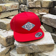 A bright red snapback cap with a diamond-shaped 'BOS' logo patch on the front, displayed on a stack of light-colored bricks against a brick wall background.