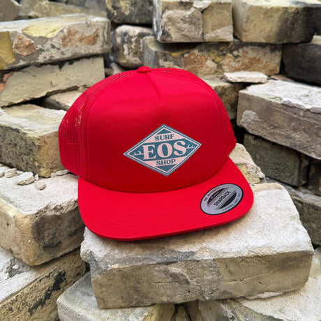A bright red snapback cap with a diamond-shaped 'BOS' logo patch on the front, displayed on a stack of light-colored bricks against a brick wall background.