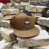A brown and tan trucker hat with a flat brim, resting on stacked stone bricks. The hat features a brown mesh back, a tan front panel, and a colorful 'EOS Surf Club' shield logo patch on the front.