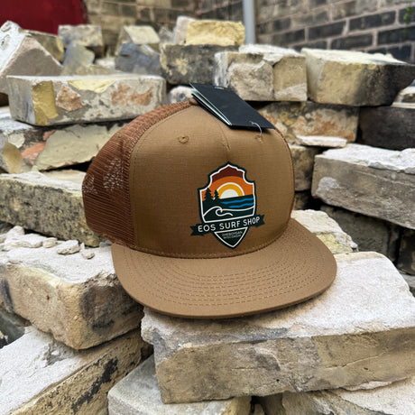 A brown and tan trucker hat with a flat brim, resting on stacked stone bricks. The hat features a brown mesh back, a tan front panel, and a colorful 'EOS Surf Club' shield logo patch on the front.