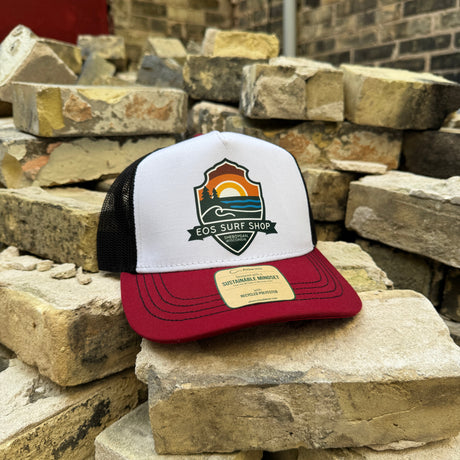 A trucker hat with a white front panel, black mesh back, and dark red brim sits on stacked stone bricks. The front panel features a colorful 'EOS SURF SHOP' shield logo, and a tan leather patch on the brim reads 'Independently Made'.