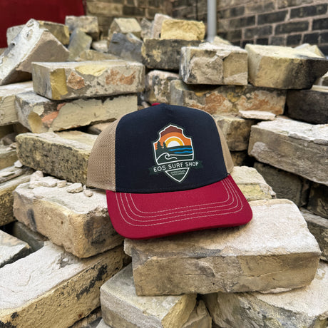 A trucker hat with a navy front panel, tan mesh back, and dark red brim sits on a pile of stacked bricks. The front panel features a colorful 'ECO SURF SHOP' logo.